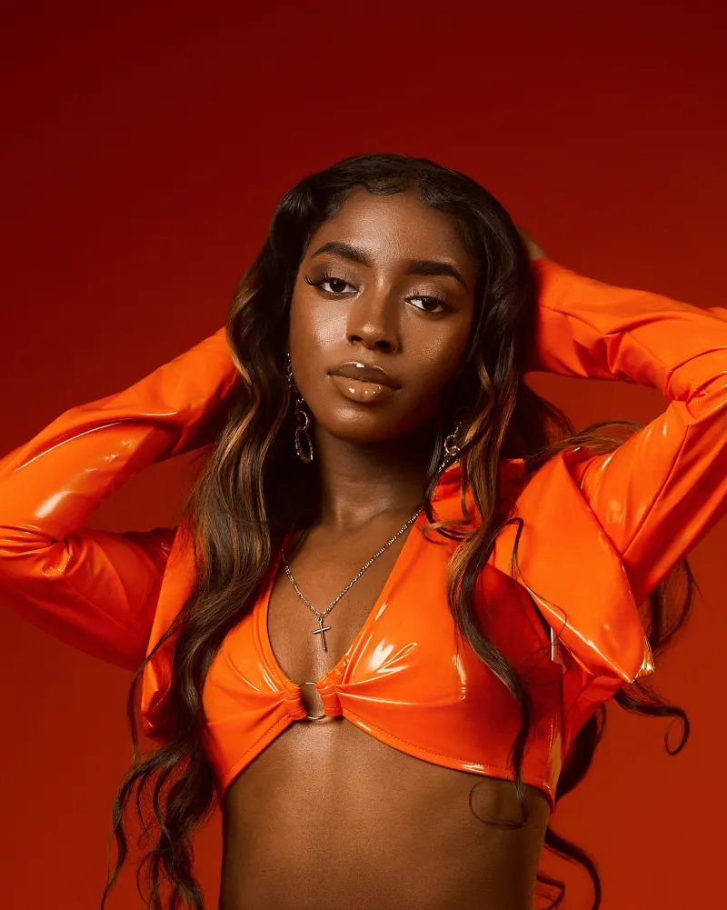 A woman in a glossy orange top with her hands in her hair against a red-orange backdrop