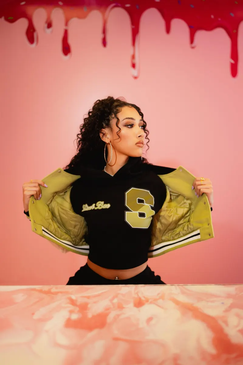 A model opening an olive varsity jacket on a pink set with dripping paint