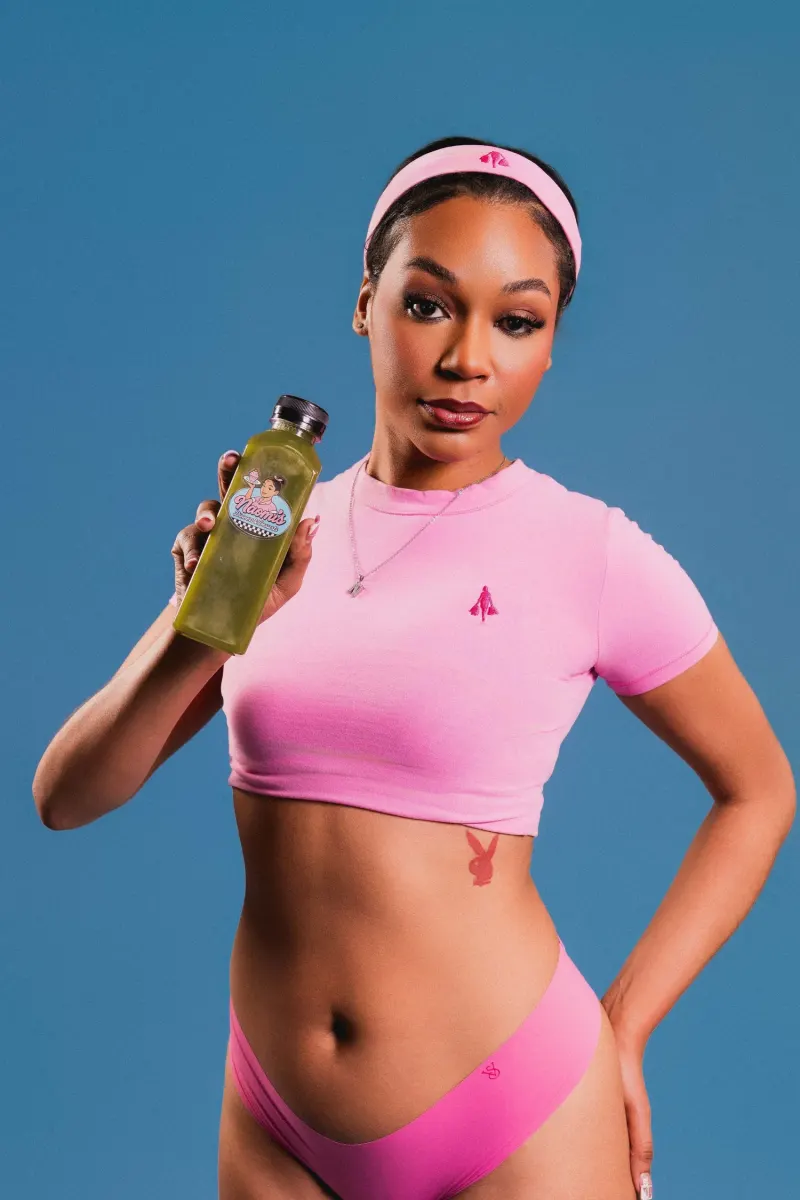 Naomi Jaee in a pink crop top and headband holding up a green juice against a blue backdrop