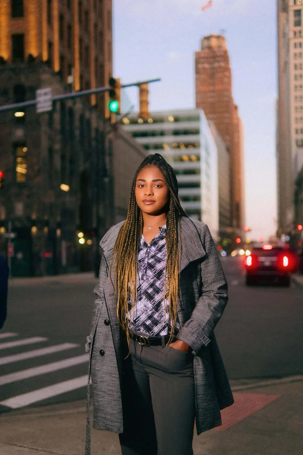 NinaJayy in a gray trench coat on a downtown Detroit street at dusk, towers and taillights behind her